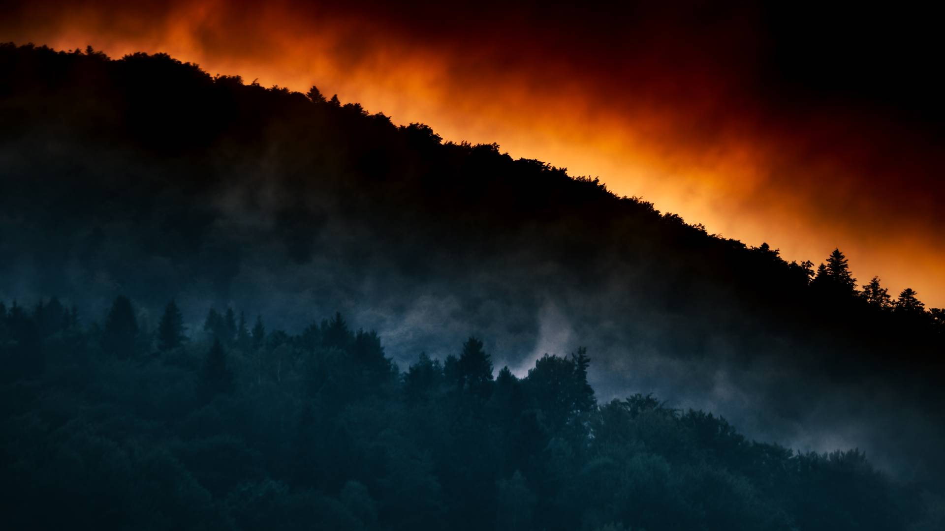 Image of hills with trees and the glow of a fire behind them